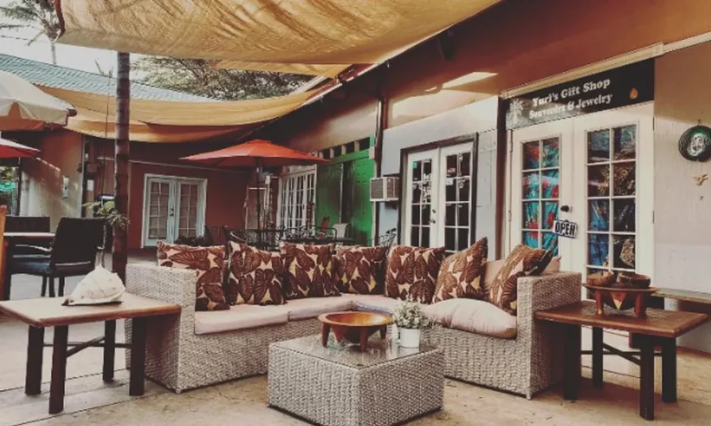 Shaded courtyard lounge with a wicker sofa, leaf-print cushions and a wooden kava bowl at Haleakava in Kihei, Maui